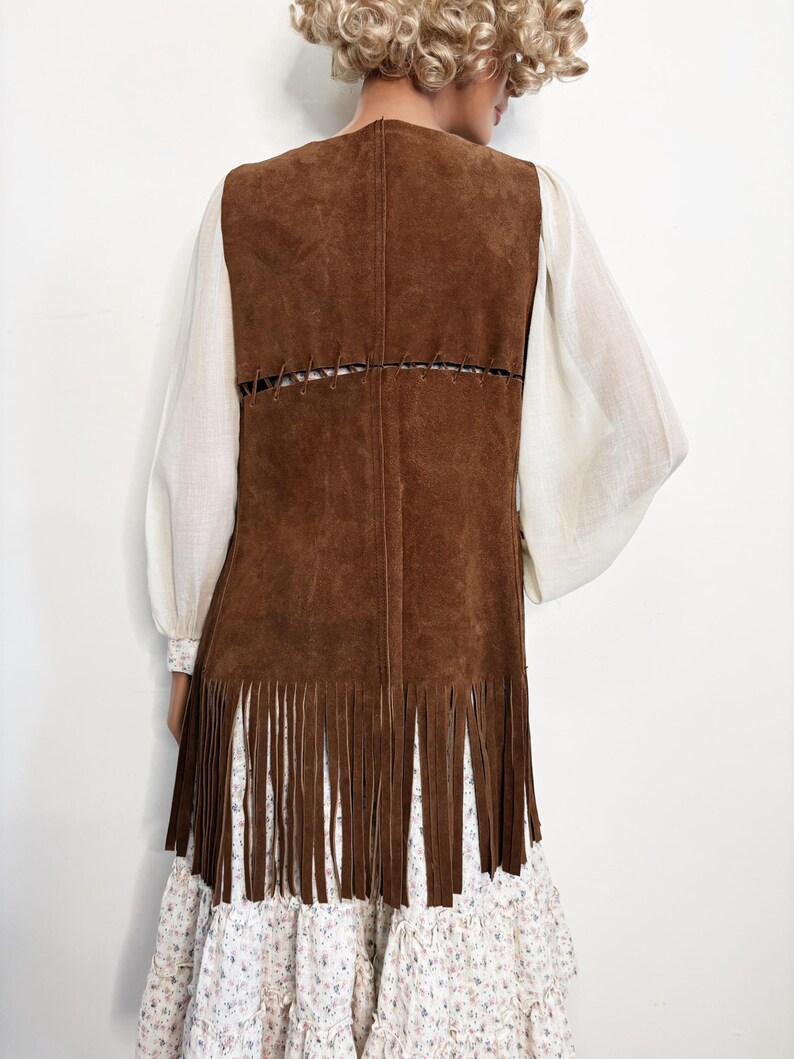 May include: A brown suede vest with long fringe at the hem. The vest has a stitched design on the back and is worn over a white blouse with billowing sleeves and a floral print dress.