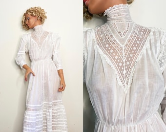 May include: A white vintage dress with a high collar and long sleeves. The dress features intricate lace detailing on the bodice and sleeves, with a tiered skirt. The dress is displayed on a mannequin.