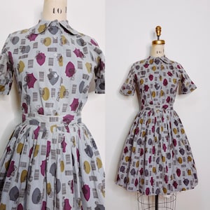 Op de afbeelding: Een vintage jurk in lichtgrijze stof, bedrukt met herhaalde patronen van paraplu's, hoeden en bagage in paars, geel en grijs. De jurk heeft een kraag, korte mouwen en een getailleerde taille met een uitlopende rok. Klassieke vintage stijl.