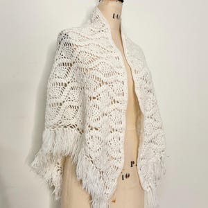 Könnte beinhalten: Cremefarbener, offener Schal mit Häkeldesign und Fransenbesatz. Der Schal ist über einer Schaufensterpuppe drapiert und zeigt sein kompliziertes Lochmuster und die strukturierte Fransen. Ein Accessoire im Vintage-Stil.