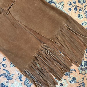 May include: A close-up of a brown suede garment with long fringe detailing. The garment appears to be a skirt or vest, with visible stitching along the seams. The fringe hangs freely, adding texture and movement to the design.