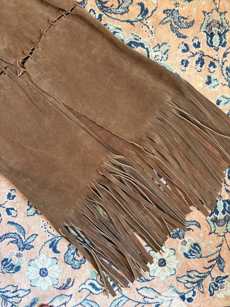 May include: A close-up of a brown suede garment with long fringe detailing. The garment appears to be a skirt or vest, with visible stitching along the seams. The fringe hangs freely, adding texture and movement to the design.