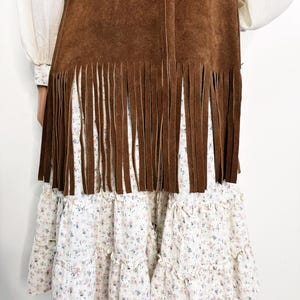May include: A brown suede vest with fringe detail over a white floral print dress. The dress has multiple ruffled tiers. The sleeves of a white blouse are visible on either side of the vest.