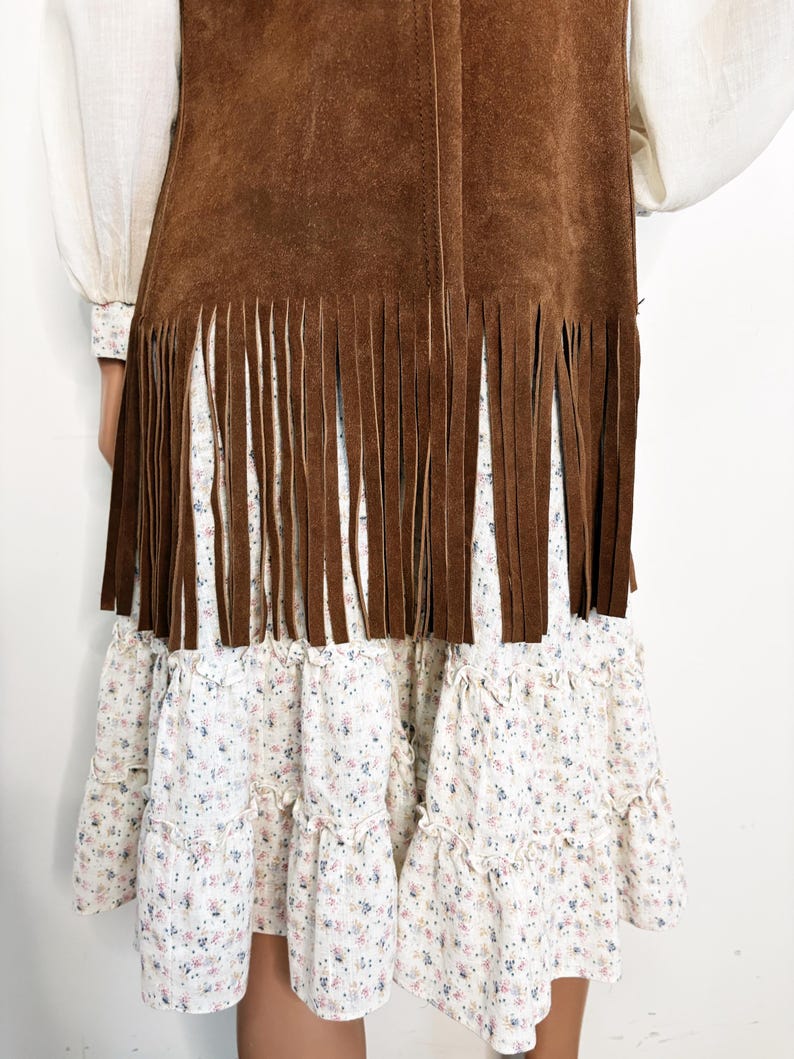 May include: A brown suede vest with fringe detail over a white floral print dress. The dress has multiple ruffled tiers. The sleeves of a white blouse are visible on either side of the vest.