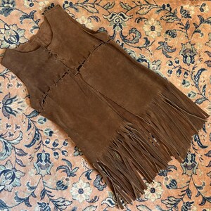 May include: A brown suede vest with a fringed hem. The vest has a sleeveless design and a V-neck. The front of the vest is detailed with decorative stitching. The vest is laying on a floral patterned rug.