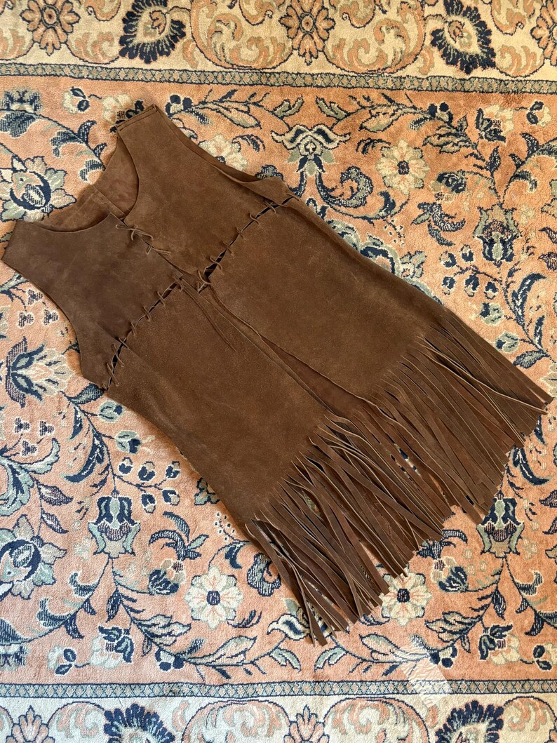 May include: A brown suede vest with a fringed hem. The vest has a sleeveless design and a V-neck. The front of the vest is detailed with decorative stitching. The vest is laying on a floral patterned rug.