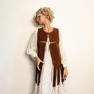 May include: A brown suede vest with long fringe, worn over a white floral print dress. The vest has a decorative cut-out design. The dress has long sleeves and a ruffled hem. The mannequin is wearing white sunglasses.