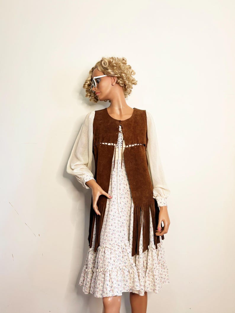 May include: A brown suede vest with long fringe, worn over a white floral print dress. The vest has a decorative cut-out design. The dress has long sleeves and a ruffled hem. The mannequin is wearing white sunglasses.