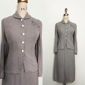 Może przedstawiać: Zestaw vintage w kratę houndstooth, prezentowany na manekinie. Dwuczęściowy zestaw zawiera kurtkę z długim rękawem z białymi guzikami i pasującą spódnicę do kolan. Garnitur jest w odcieniach brązu i bieli.
