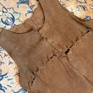 May include: Brown suede vest with a sleeveless design. The vest features decorative stitching and ties along the front. The garment is a medium brown color.