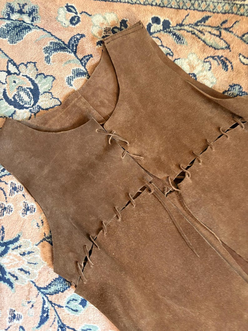 May include: Brown suede vest with a sleeveless design. The vest features decorative stitching and ties along the front. The garment is a medium brown color.
