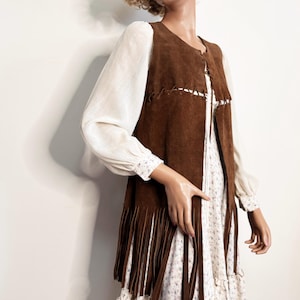 May include: A brown suede vest with long fringe, worn over a white floral print dress. The vest has a stitched design along the edges. The dress has long sleeves and a ruffled hem. The mannequin is wearing white sunglasses.