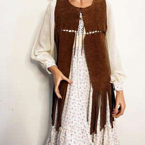 May include: A brown suede vest with fringe detail, worn over a floral print dress with long sleeves. The dress has a ruffled hem and a white background with small pink and blue flowers. The vest has a round neckline and a lace-up closure.