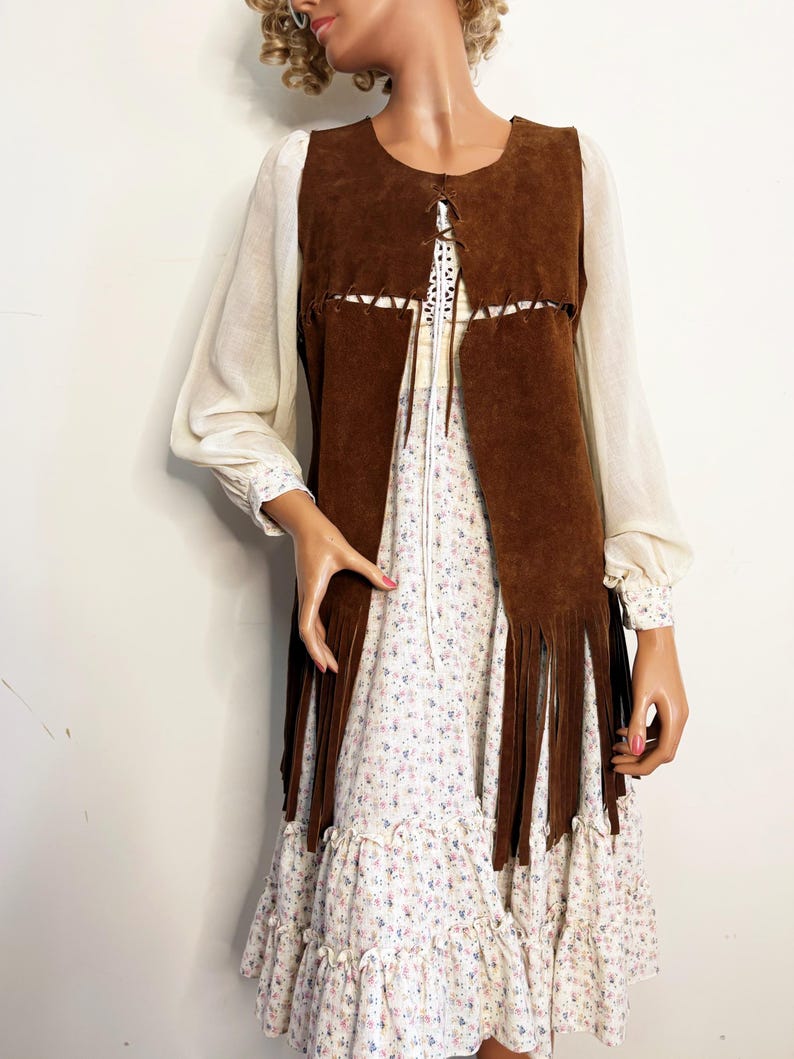 May include: A brown suede vest with fringe detail, worn over a floral print dress with long sleeves. The dress has a ruffled hem and a white background with small pink and blue flowers. The vest has a round neckline and a lace-up closure.