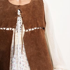 May include: A brown suede vest with a lace-up front detail. The vest is layered over a white crochet top and a floral print dress. The sleeves of a cream-colored blouse are visible on either side of the vest.