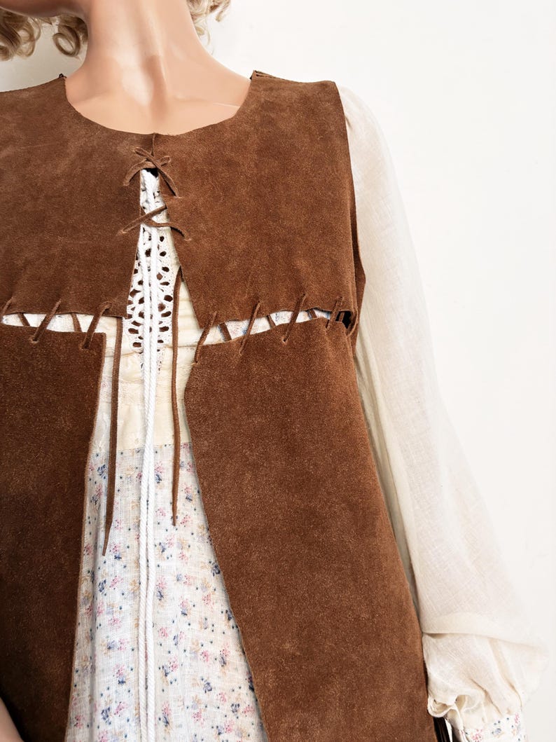 May include: A brown suede vest with a lace-up front detail. The vest is layered over a white crochet top and a floral print dress. The sleeves of a cream-colored blouse are visible on either side of the vest.