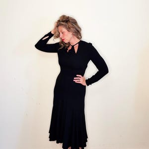 May include: A black long-sleeved midi dress featuring a fitted bodice, a flared skirt, and a unique cutout detail at the neckline. The sleeves have small slits at the cuffs, and the dress is paired with black pointed-toe shoes.
