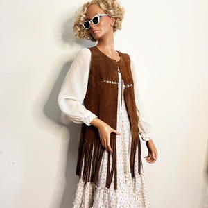 May include: A mannequin wearing a brown suede vest with long fringe, over a white floral print dress. The vest has decorative stitching and the dress has long sleeves. The mannequin is also wearing white sunglasses.