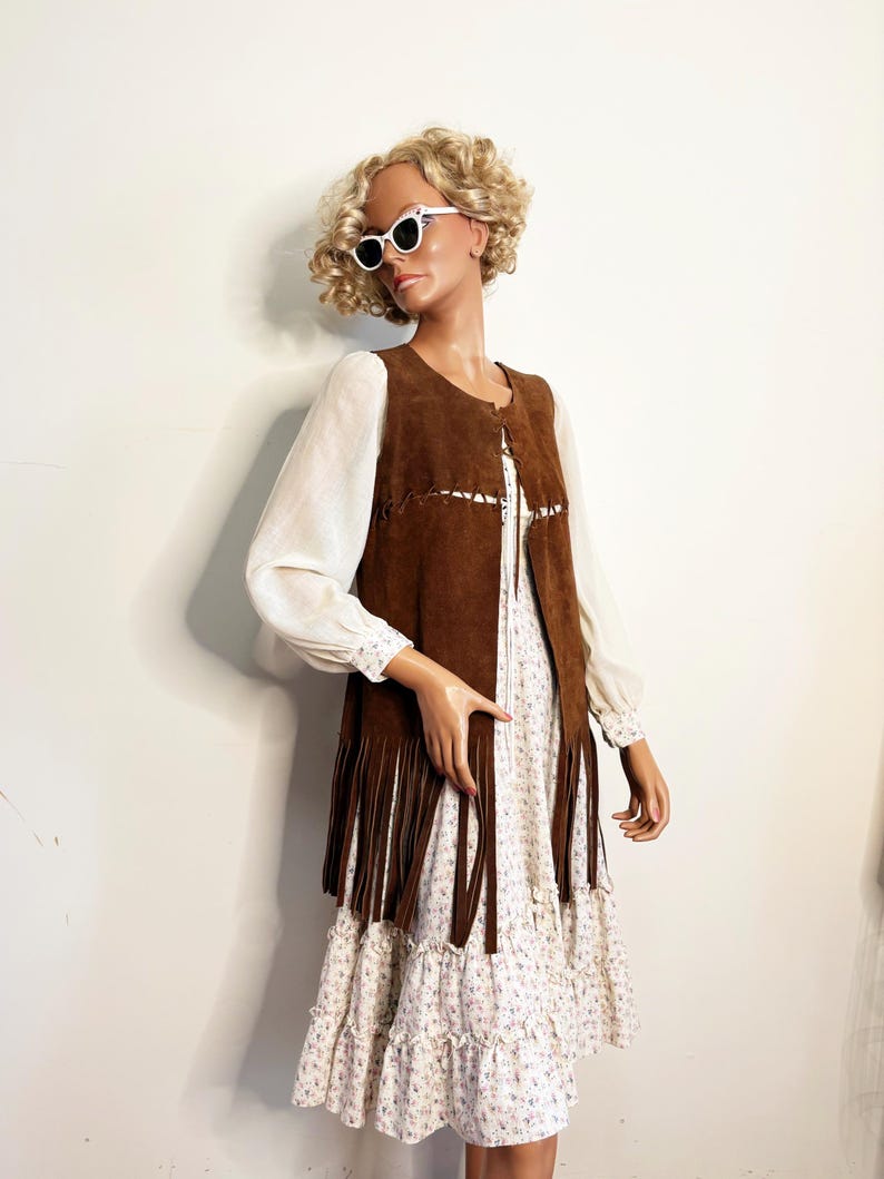 May include: A mannequin wearing a brown suede vest with long fringe, over a white floral print dress. The vest has decorative stitching and the dress has long sleeves. The mannequin is also wearing white sunglasses.