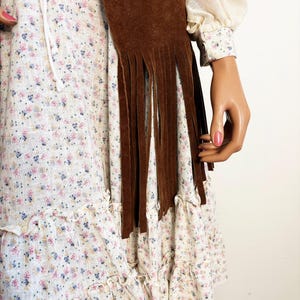 May include: A brown suede fringed scarf is draped over a white floral print dress. The dress has a ruffled hem and a drawstring neckline. The sleeves are long and the cuffs are patterned with small flowers.