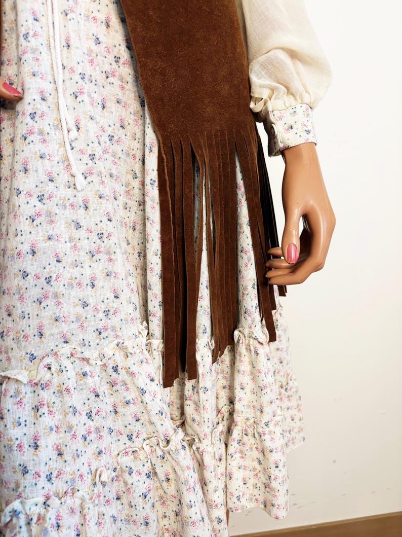 May include: A brown suede fringed scarf is draped over a white floral print dress. The dress has a ruffled hem and a drawstring neckline. The sleeves are long and the cuffs are patterned with small flowers.