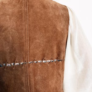 May include: A brown suede vest with visible stitching details. The vest has a vertical seam down the back and horizontal stitching across the lower back. A white long-sleeved shirt is visible on the right side of the vest.