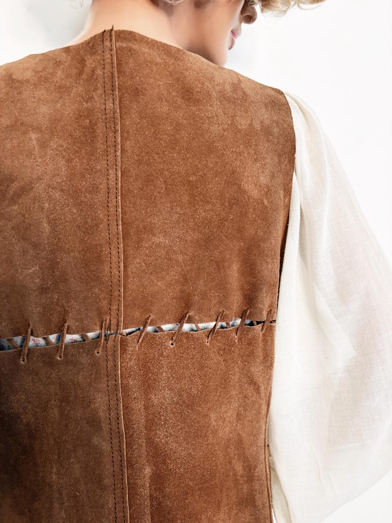 May include: A brown suede vest with visible stitching details. The vest has a vertical seam down the back and horizontal stitching across the lower back. A white long-sleeved shirt is visible on the right side of the vest.