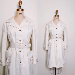May include: A white long-sleeved trench coat with a belt and large pockets. The coat is on a white mannequin.
