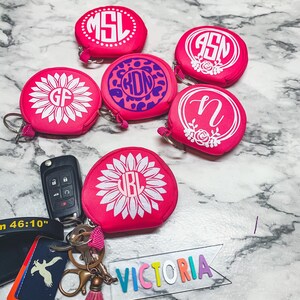 May include: Six pink round zippered pouches with white and purple monograms and floral designs. The pouches are attached to key rings. The word "VICTORIA" is spelled out in rainbow letters on a clear plastic banner.
