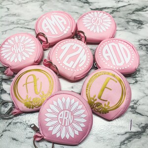 May include: Eight pink zippered pouches with various white and gold monograms and sunflower designs. The pouches are decorated with the letters "AMA", "EZP", "KWE", "12M", "ADD", "A", "E", and "ARN".