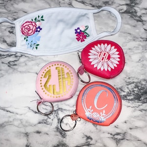 May include: Three pink and coral zippered pouches with gold and silver lettering and floral designs. Each pouch has a silver key ring attached. The pouches are on a white marble surface.