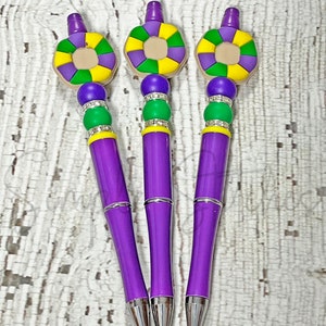 May include: Three purple pens with silver accents. Each pen features a decorative top with a purple, green, and yellow design, and a beaded section with purple and green beads. The pens are arranged on a white, textured surface.