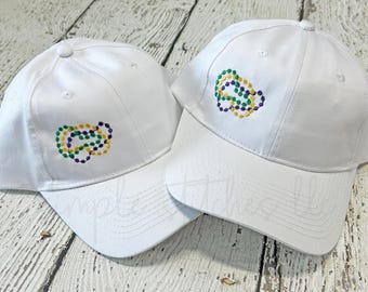 May include: Two white baseball caps with embroidered Mardi Gras bead designs in purple, green, and yellow. The caps have a curved brim and a button on top. The caps are displayed on a weathered, light-colored wooden surface.