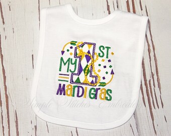 May include: White baby bib embroidered with "My 1st Mardi Gras" in green, purple, and gold. The number "1" is decorated with a harlequin pattern and a gold bead necklace. Festive confetti accents the design.