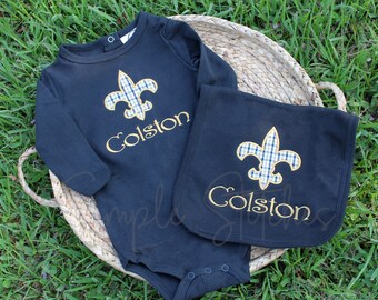 May include: A navy blue baby onesie and bib set. Each item features a gold fleur-de-lis with a blue and white plaid pattern. The name "Colston" is embroidered in gold script on both pieces. The set is displayed on a woven basket.