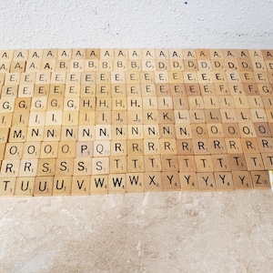 May include: Wooden Scrabble tiles arranged on a light-colored surface. The tiles display letters of the alphabet, including multiple instances of common letters like 'E' and 'A'. A yellow tile with the letter 'G' is visible.