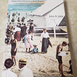 May include: A vintage book cover titled "THE TOURIST" with the subtitle "Travel in Twentieth-Century North America." The cover features a sepia-toned photograph of people on a beach. The author's name, John A. Jakle, is also visible.