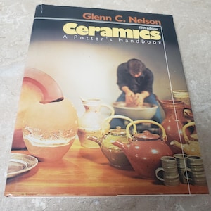 May include: A book titled "Ceramics: A Potter's Handbook" by Glenn C. Nelson, fifth edition. The cover features a potter at work, alongside various ceramic pieces, including teapots, mugs, and vases, all displayed on a wooden surface.