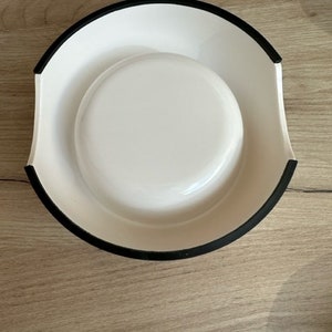 May include: White ceramic pet bowl with a black rim. The bowl has a raised lip around the edge.