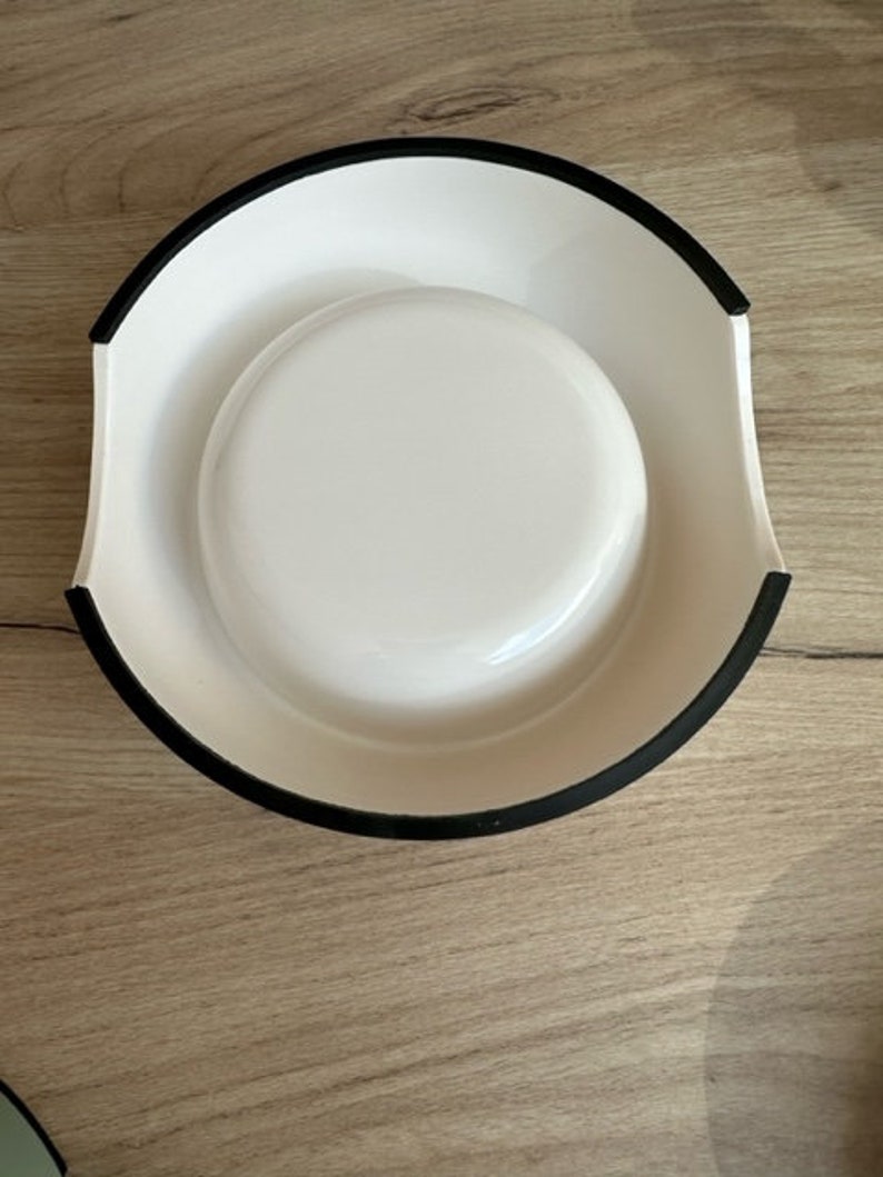 May include: White ceramic pet bowl with a black rim. The bowl has a raised lip around the edge.