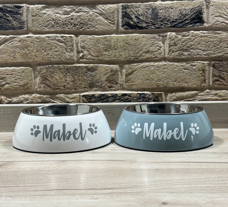 May include: Two stainless steel pet bowls with a white and gray finish. The bowls are personalized with the name "Mabel" and paw prints.