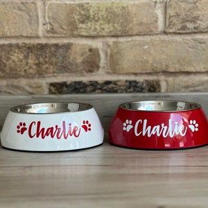 May include: Two personalized pet bowls, one white and one red, with the name "Charlie" and paw prints in red on the white bowl and in white on the red bowl.