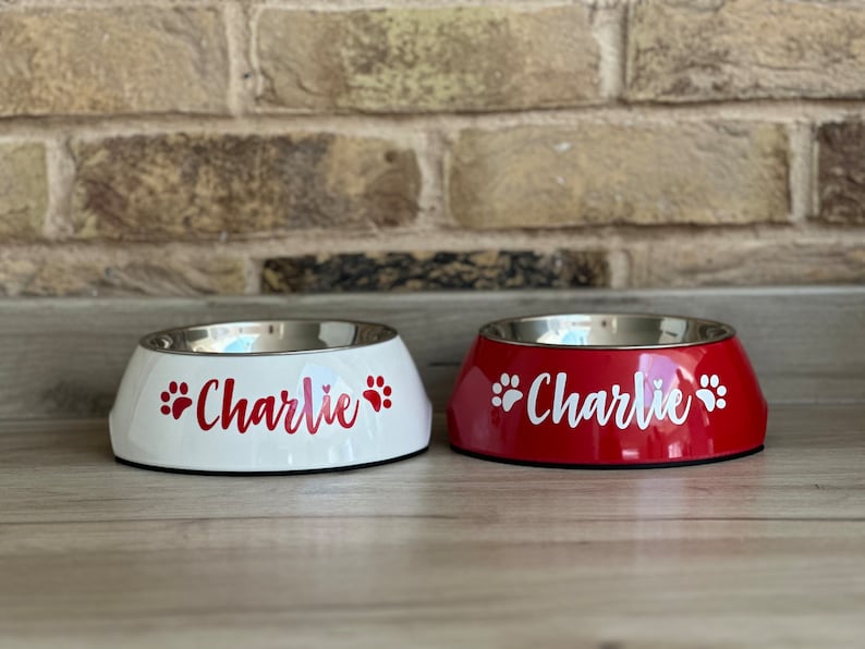 May include: Two personalized pet bowls, one white and one red, with the name "Charlie" and paw prints in red on the white bowl and in white on the red bowl.