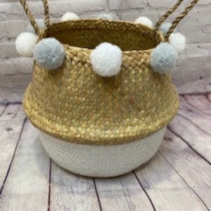 May include: A natural woven basket with white and grey pom poms around the top. The basket has a white base and natural woven top.