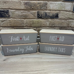 May include: Four grey storage boxes with wooden lids. Two boxes are labelled "First + Aid" and two are labelled "Laundry Tabs" in white script. The boxes are stacked on a wooden surface, with a brick wall in the background.