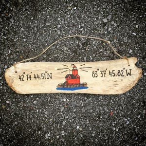 May include: A wooden sign with a painted lighthouse and coordinates: 42&deg; 14' 44.51" N 83&deg; 37' 45.82" W.