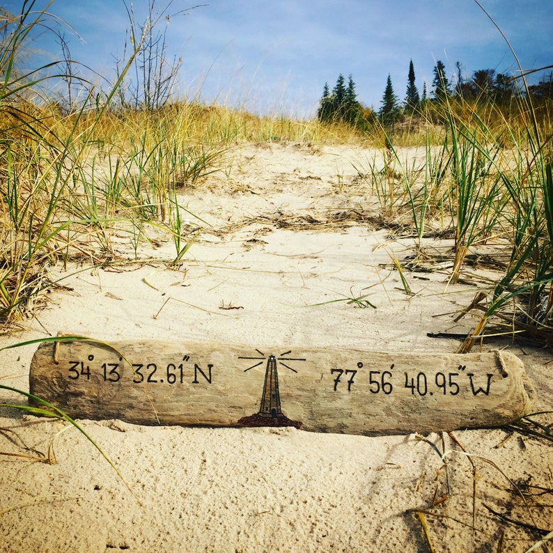 May include: A piece of driftwood with coordinates and a lighthouse drawing carved into it. The coordinates are 34&deg; 13' 32.61" N and 77&deg; 56' 40.95" W. The driftwood is lying on a sandy beach with green grass in the background.