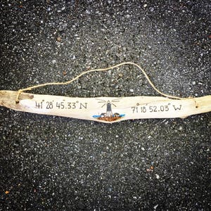 May include: A piece of driftwood with a lighthouse drawing and the coordinates "41&deg; 28' 45.33" N 71&deg; 18' 52.03" W" written on it.