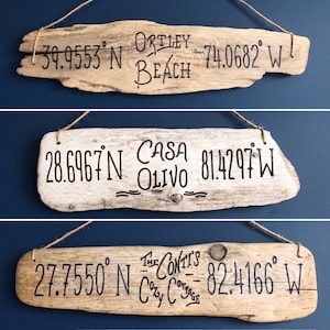 May include: Three rectangular driftwood signs hanging from twine. The top sign reads Ortley Beach with coordinates 39.9553° N 74.0682° W. The middle sign reads Casa Olivo with 28.6967° N 81.4297° W. The bottom sign reads The Conti's Cozy Cottage 27.7550° N 82.4166° W.