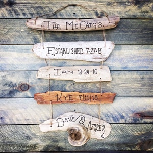 May include: Five rustic wooden signs with handwritten text. The top sign reads "The McCaiG's". The second sign reads "Established 7-27-13". The third sign reads "Ian 12-24-16". The fourth sign reads "Kye 7/11/18". The bottom sign reads "Dave & Amber".